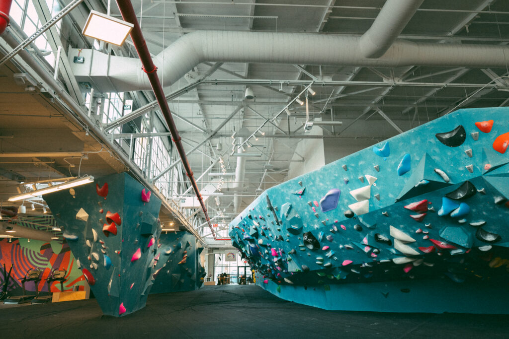 A bright indoor climbing gym with large blue bouldering walls covered in colorful holds, high ceilings, exposed pipes, and natural light streaming in—a perfect spot to follow a weekly training routine for climbers.