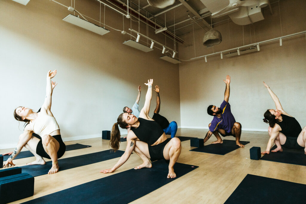 Five people practice yoga in a bright studio, performing a seated twist pose on black mats with yoga blocks nearby. This session is part of a weekly training routine for climbers, helping the group improve focus, balance, and flexibility.