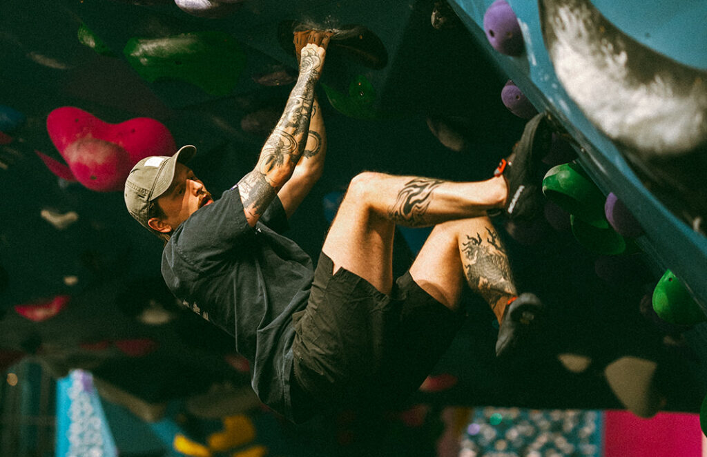 A person with tattoos climbs an indoor bouldering wall at a fitness-focused climbing gym, hanging from large holds with both hands and feet. The gym features colorful grips and ample padding below, offering an engaging alternative to traditional gyms.
