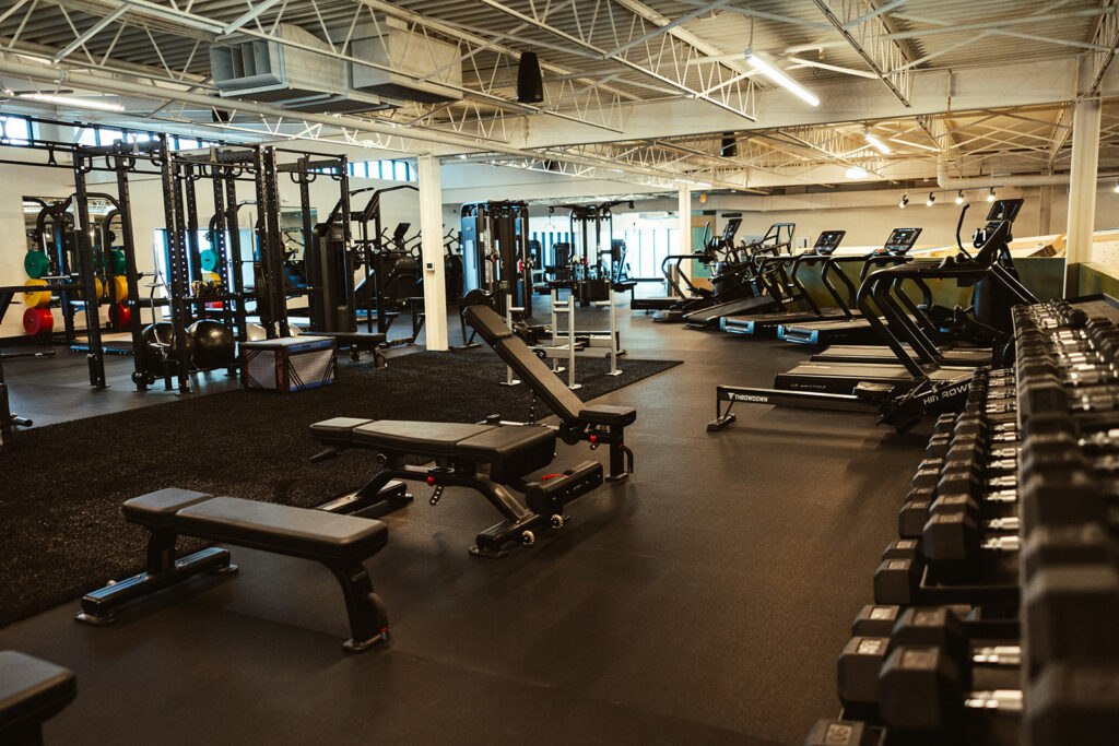 Spacious gym with weight benches, dumbbells, squat racks, and cardio machines like treadmills and ellipticals. Large windows provide natural light, creating an inviting atmosphere for anyone focused on fitness. The floor is covered with black mats, offering both comfort and durability—not unlike the practical surfaces often found in traditional gyms.