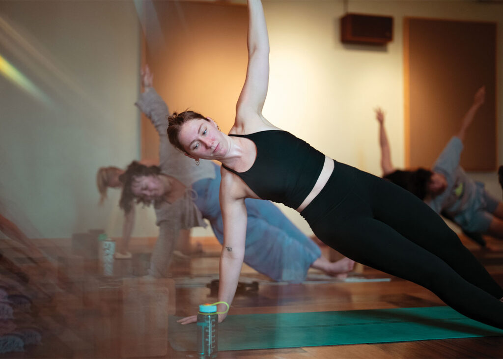 A woman in black athletic wear performs a side plank in a yoga class, demonstrating the functional benefits often associated with Pilates, while other participants are blurred in the background.