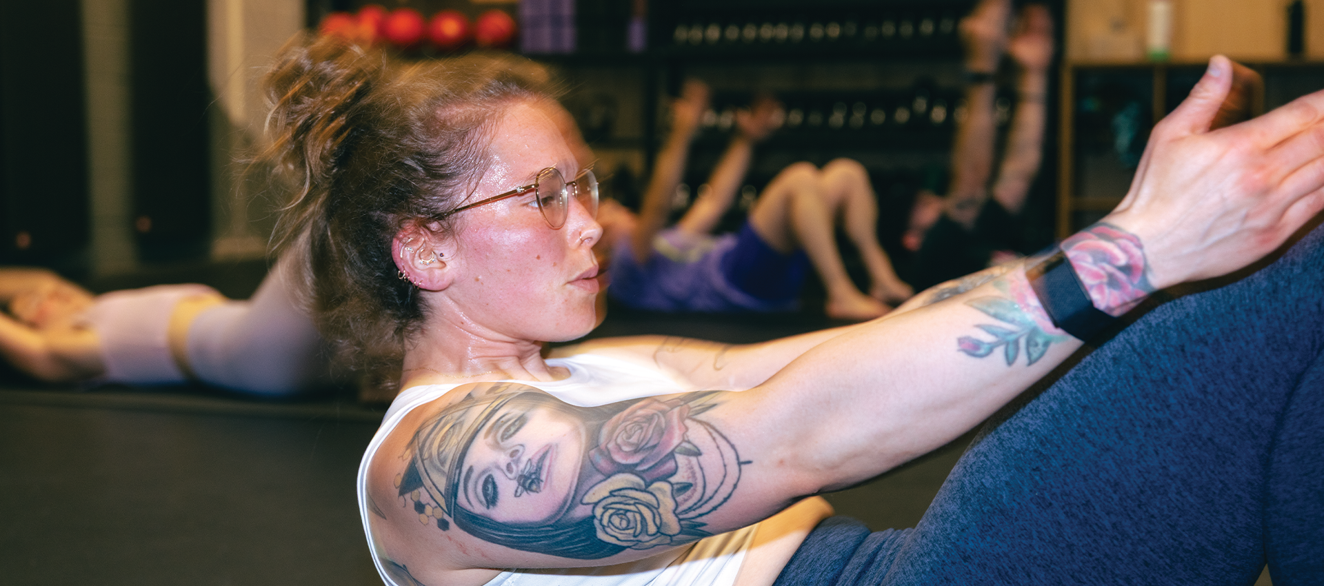 A woman with glasses and arm tattoos does a Pilates fitness exercise on a gym floor, enjoying the functional benefits of her workout, with others working out in the background.
