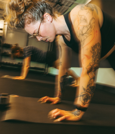 A person with tattoos and glasses does a plank exercise on a mat in a gym, showcasing the functional benefits of Pilates, with another person blurred in the background, suggesting motion and intensity.