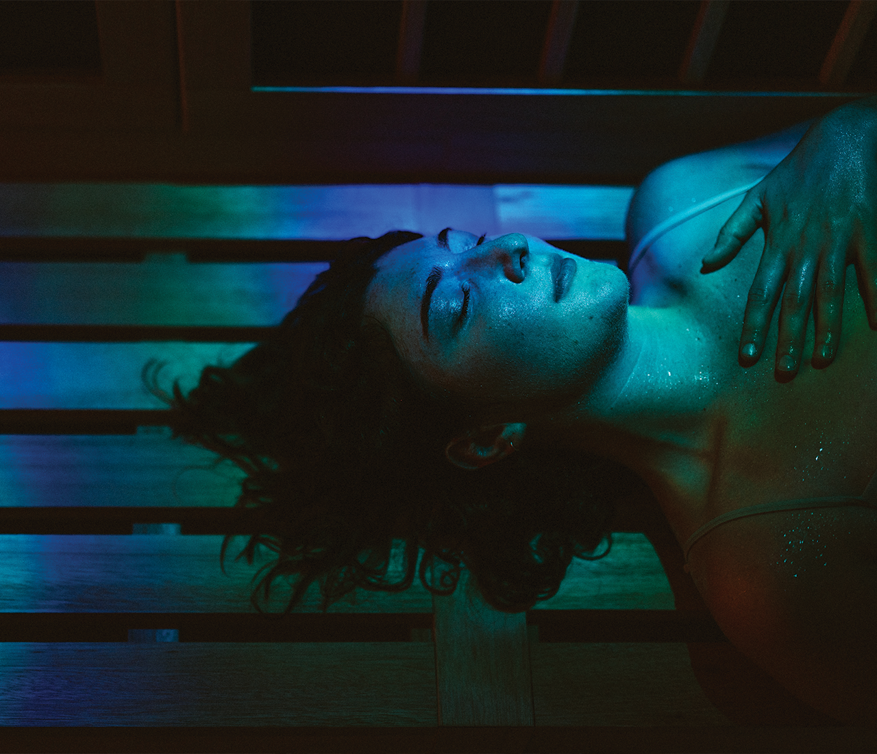 A person lies on a wooden surface under blue and green light, eyes closed, with one hand resting on their chest, appearing calm and relaxed—enjoying the tranquil amenities of Austin Bouldering Project.