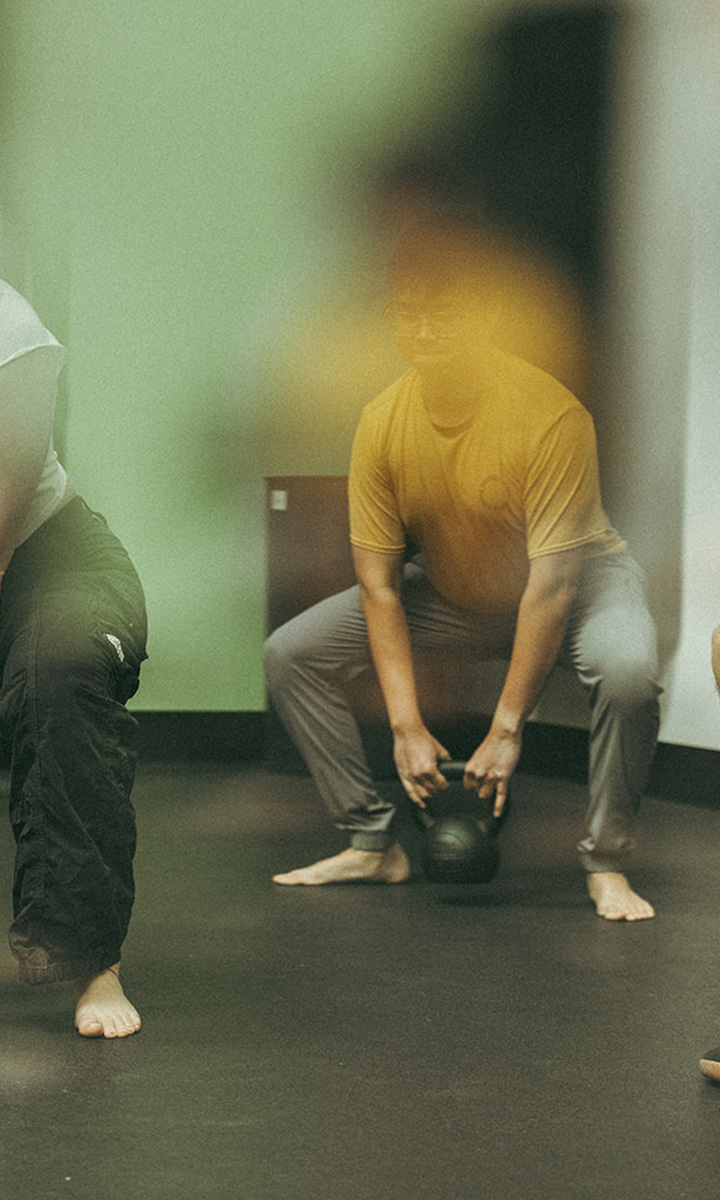 A person in a yellow shirt and gray pants is squatting barefoot indoors, holding a kettlebell, as part of the Summer Streak Challenge. Another person is partially visible on the left, also barefoot and squatting. The image has a soft, slightly blurred effect, capturing the energy of this Seattle fitness session.