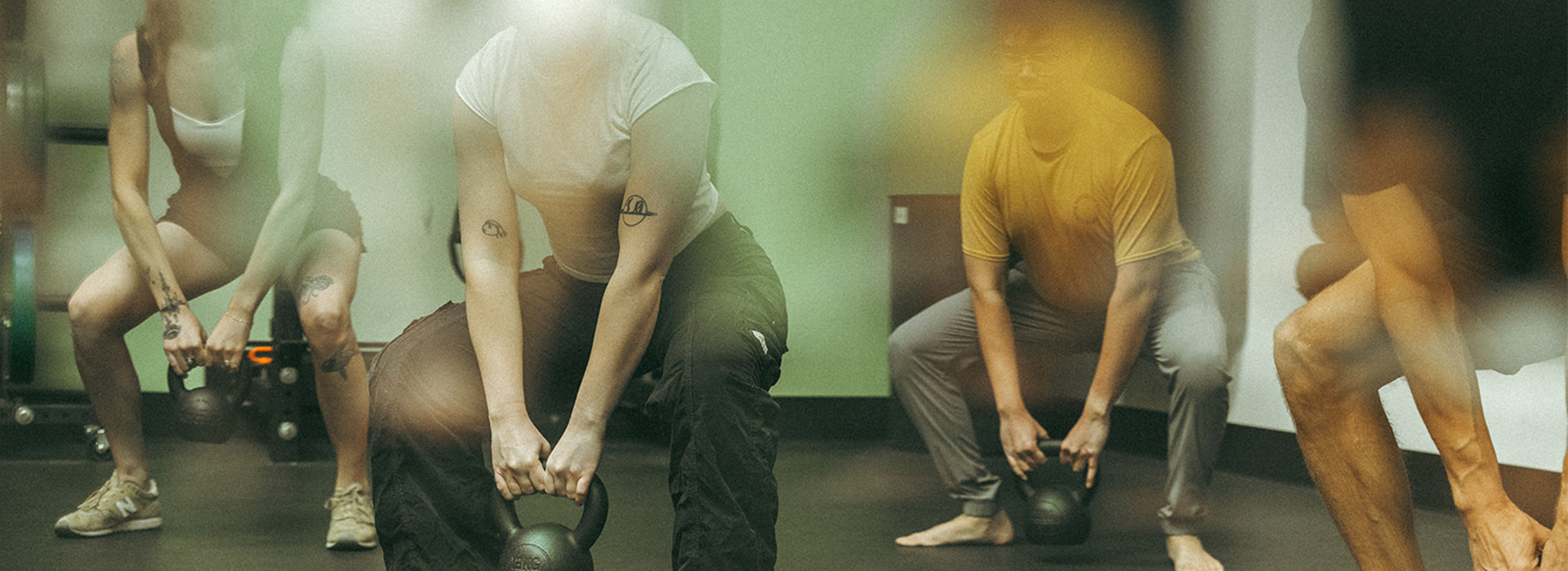 A group of people in athletic wear are exercising indoors in Seattle, holding kettlebells and bending forward at the waist, focusing on their workout as part of the Summer Streak Challenge.