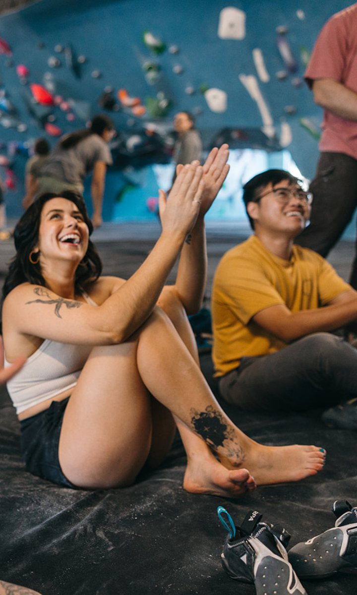 A woman sits cross-legged and clapping, smiling, next to a man in glasses at an indoor climbing gym in Tempe. Both are barefoot with climbing shoes nearby as they enjoy their First Visit Free, while people climb in the background.