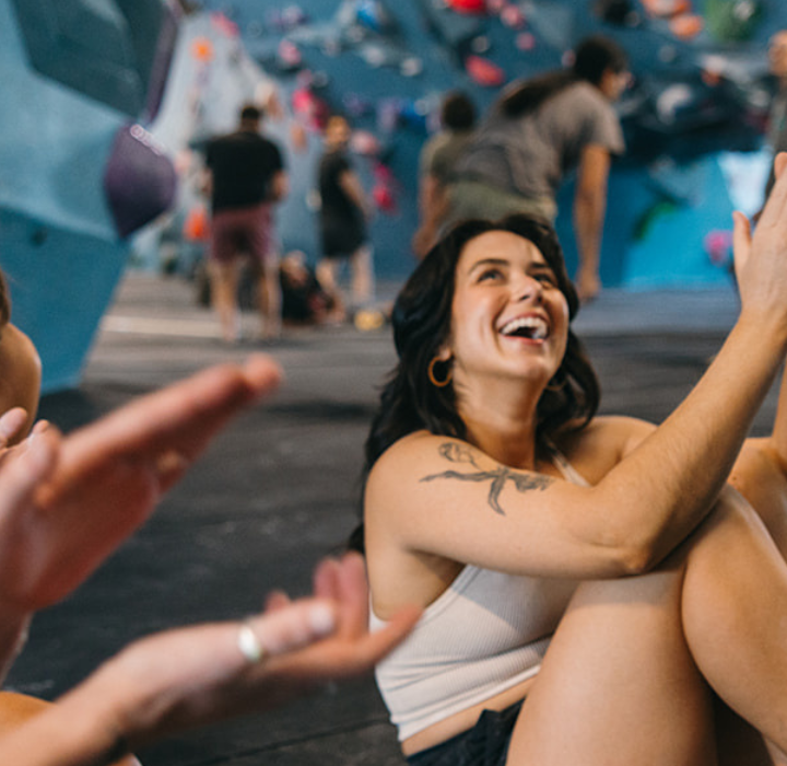 A group of young adults sit on a gym floor in Tempe, smiling and clapping, with colorful indoor climbing walls in the background—perfect for anyone looking to try something new, especially with a First Visit Free offer.
