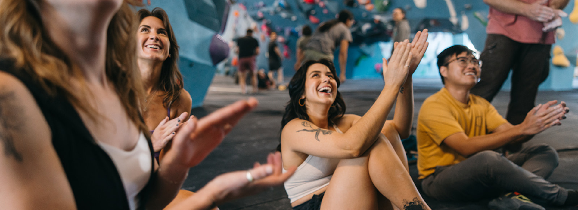 A group of young adults sit on a gym floor in Tempe, smiling and clapping, with colorful indoor climbing walls in the background—perfect for anyone looking to try something new, especially with a First Visit Free offer.