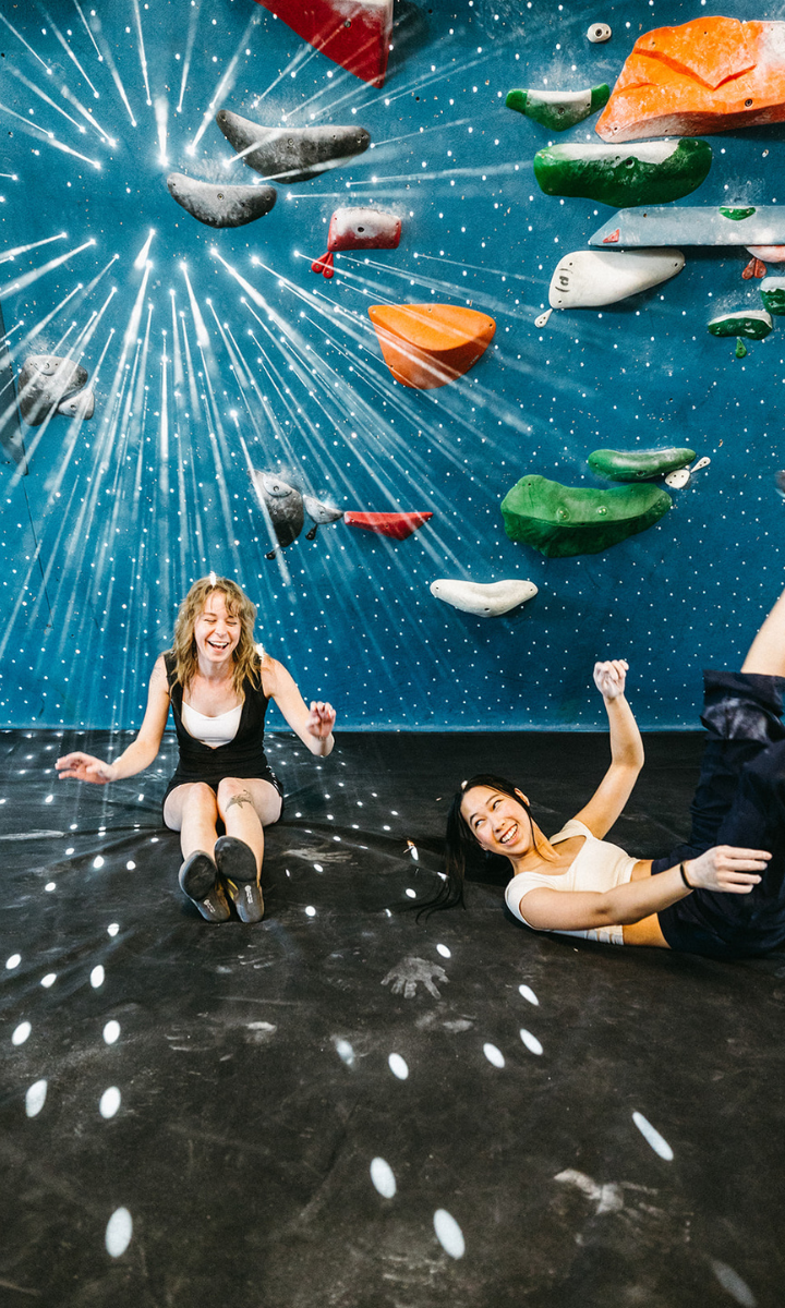 Two women laugh and sit on the floor of an indoor climbing gym in Tempe, surrounded by colorful holds and starburst light patterns projected on the blue wall and ground—soaking in the excitement of their first visit free.