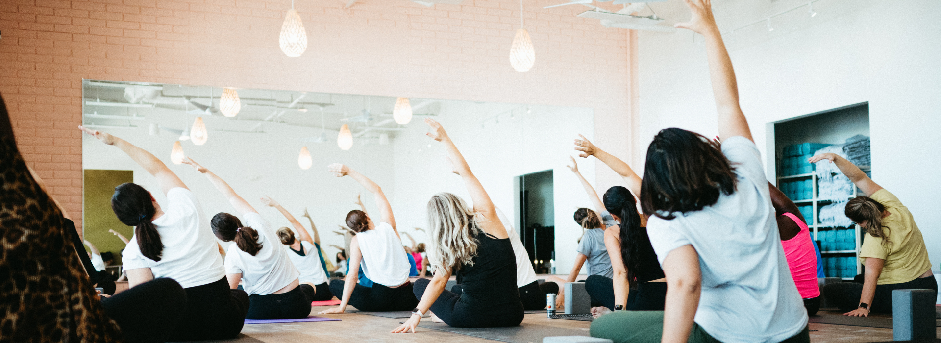 A group of people in a bright Tempe studio sit on yoga mats facing a mirror, all stretching their left arms overhead in unison during a yoga or fitness class. First Visit Free.