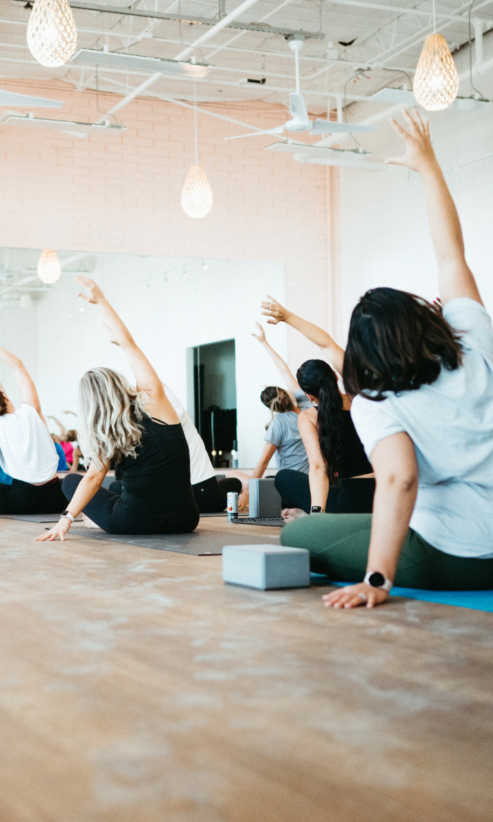 A group of people in workout clothes sits on yoga mats in a Tempe studio, stretching their arms overhead during a yoga class. The room has wooden floors and soft lighting, creating a welcoming atmosphere—perfect for newcomers, with First Visit Free offers available.