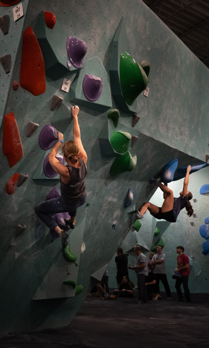 Two climbers ascend an indoor bouldering wall with colorful red, green, and purple holds in Minneapolis while several people look on. Perfect for a first visit free—watch their progress before heading to the confirmation page to reserve your spot!.