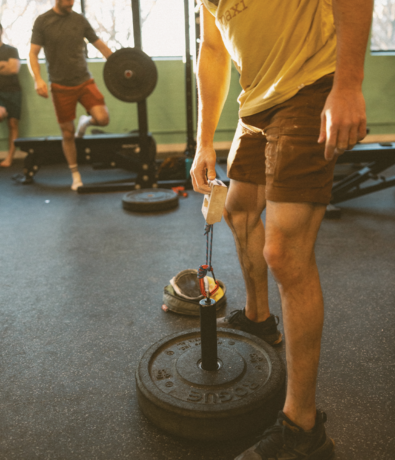 A person in shorts and a yellow shirt grips a handle attached to stacked weight plates at a gym, preparing for a strength exercise as part of a weekly training routine for climbers. Another person stands in the background near gym equipment.