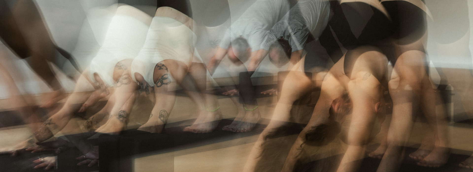 A blurred image of several people in a Minneapolis yoga studio performing the downward dog pose, creating a sense of motion. Some have tattoos and are wearing athletic clothing. A perfect glimpse into your First Visit Free experience.
