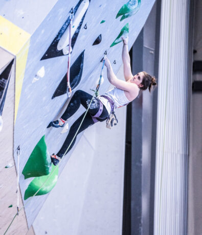 A woman climbs an indoor rock wall in Brooklyn, gripping green holds and secured by a harness and ropes. She wears a white tank top, black leggings, and climbing shoes, benefiting from personal coaching to hone her skills.