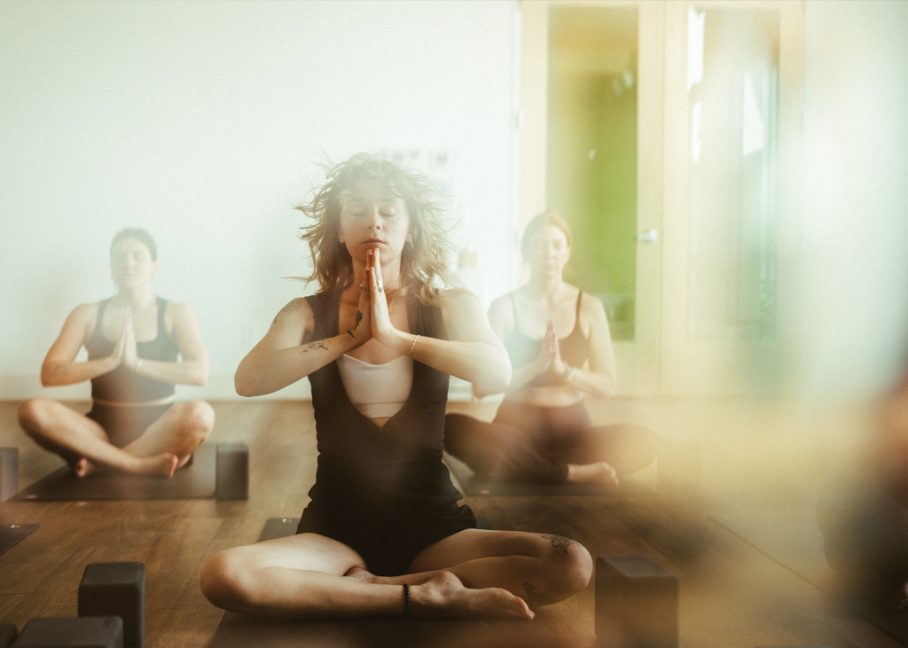 Three people sit cross-legged on yoga mats indoors, eyes closed, hands pressed together in front of their chests, practicing meditation in a calm, softly-lit room—a tranquil moment from one of the Signature Yoga Classes at Bouldering Project.