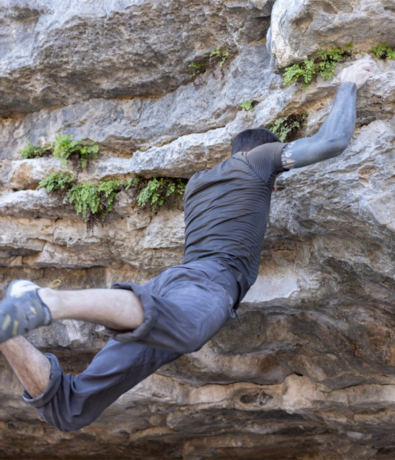 A person wearing dark clothes climbs a rocky surface—perhaps one of the best places for Austin climbing—reaching with one arm while their legs dangle. Small green plants grow in crevices of the rock.