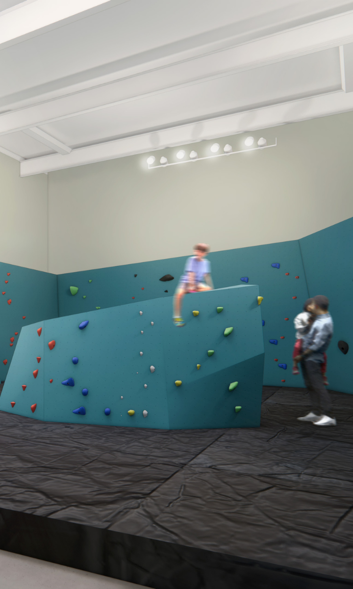 Two people in a modern DC climbing gym with padded floors, colorful climbing holds on blue walls, and bright overhead lighting. One person is climbing while another stands nearby.