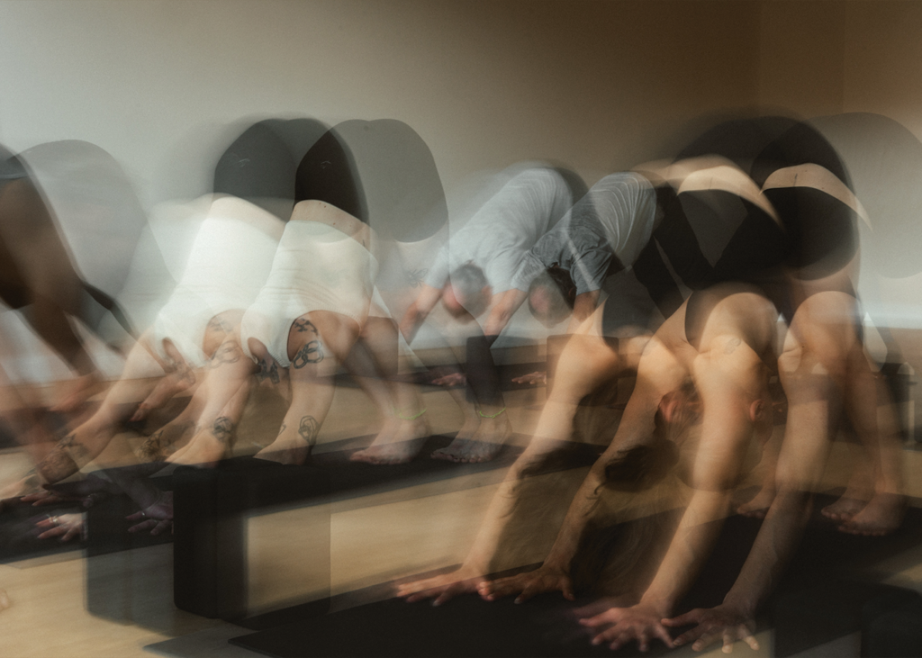 A group of people practicing yoga at the Bouldering Project, captured with a motion blur effect as they move into a downward dog pose on yoga mats in a studio that also offers bouldering and yoga classes.