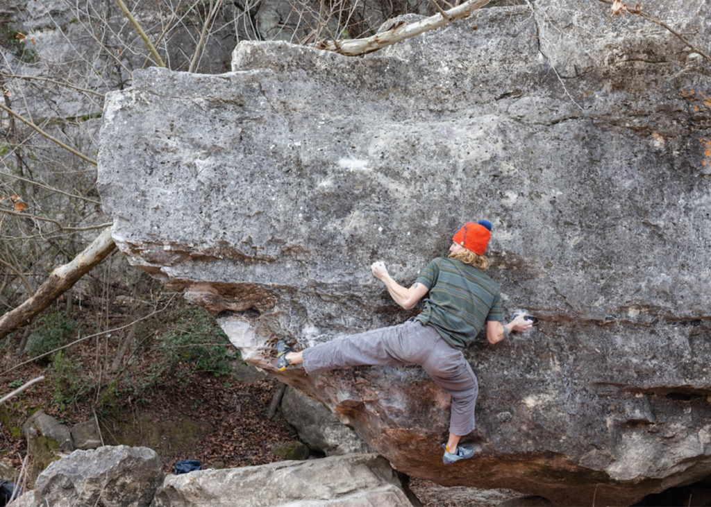 A person wearing a red beanie climbs a large, overhanging rock outdoors—an exciting scene that captures the spirit of rock climbing Austin has to offer. They grip the rock face with both hands and balance expertly on small footholds, surrounded by leafless trees and rugged rocks—hallmarks of some of the best places Austin climbers love to explore.