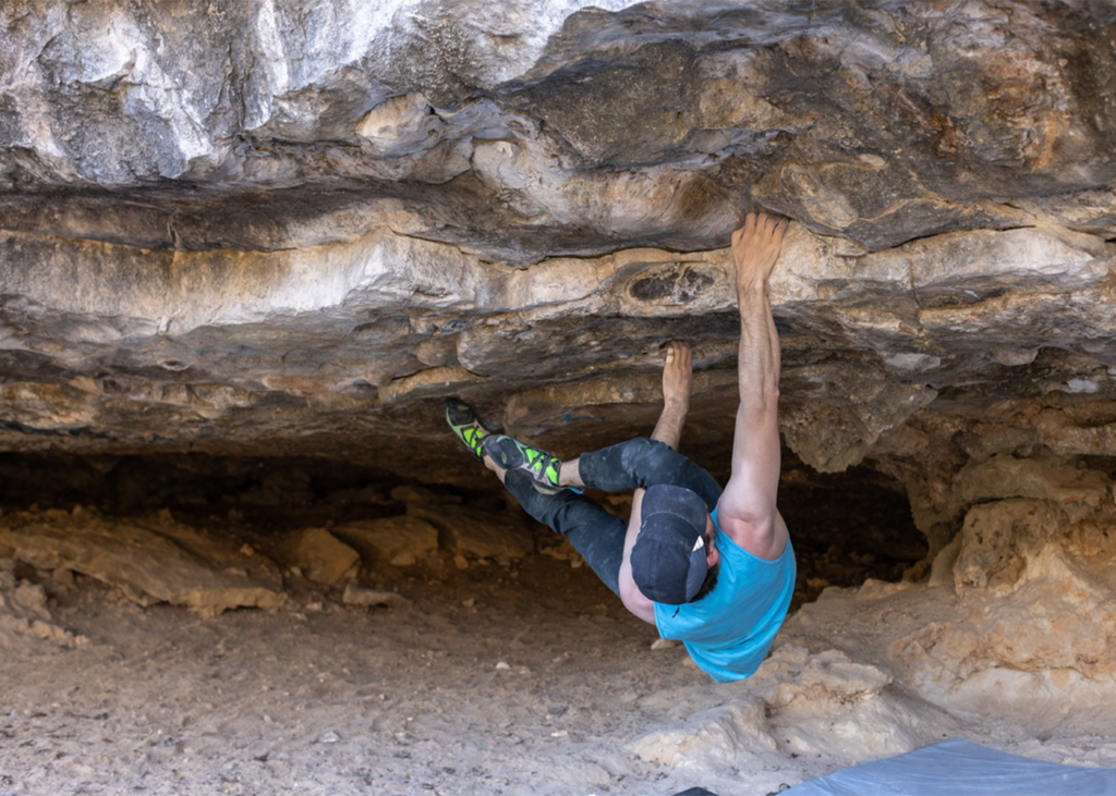 A person in a blue tank top and cap is bouldering, gripping the underside of a rocky overhang with both hands and feet, wearing climbing shoes. This scene captures the thrill of rock climbing Austin offers, showcasing one of the best places for Austin climbing enthusiasts to challenge themselves.