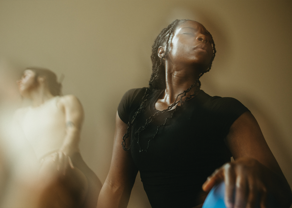 Two people, one in focus and one blurred in the background, appear to be stretching or engaging in signature yoga poses in soft light. The person in front, with braided hair and a black shirt, has their eyes closed and an expressive, serene facial expression.