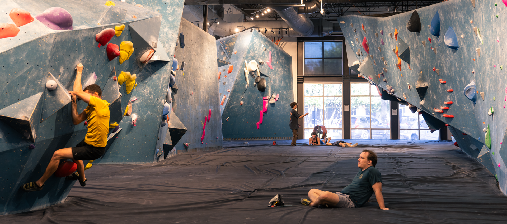 Several people are bouldering inside a climbing gym with blue walls and colored holds. One person climbs on the left while others sit or stand on the padded floor, and large windows let in daylight. It’s a welcoming space to start rock climbing and experience the growing rock climbing Austin community.