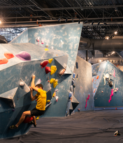 A person in a yellow shirt is bouldering on an indoor climbing wall with various colored holds at an Austin rock climbing gym; more climbing walls are visible in the background.