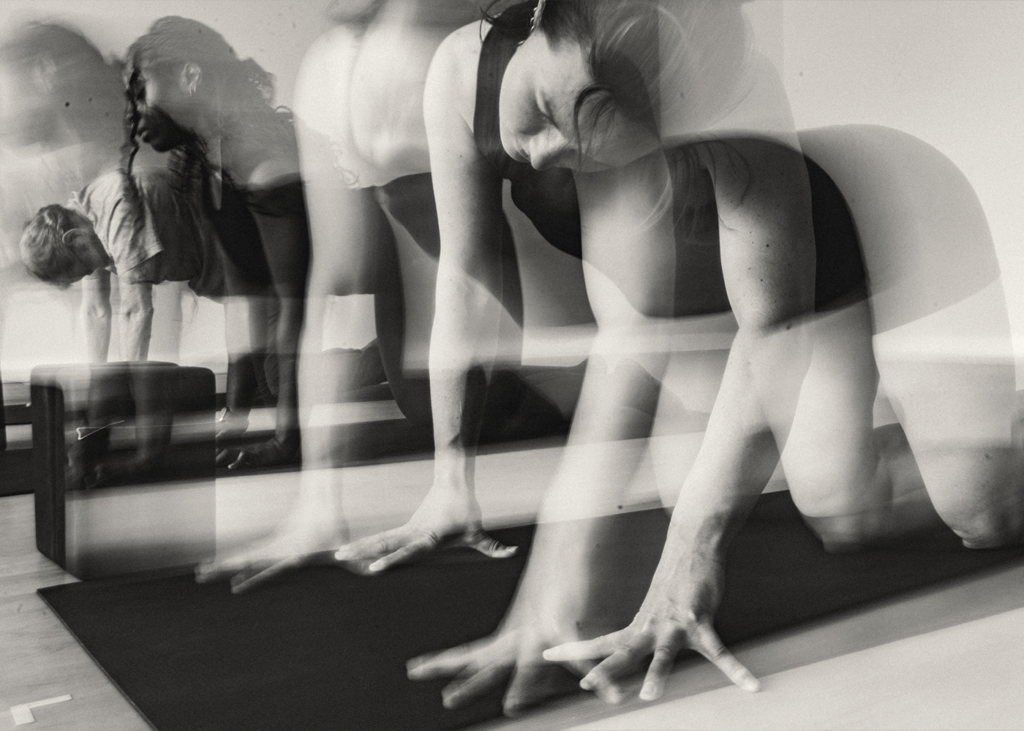 Black and white photo showing a blurred, overlapping image of people doing yoga or pilates on mats at the Bouldering Project, emphasizing motion and fluidity in their signature yoga poses.