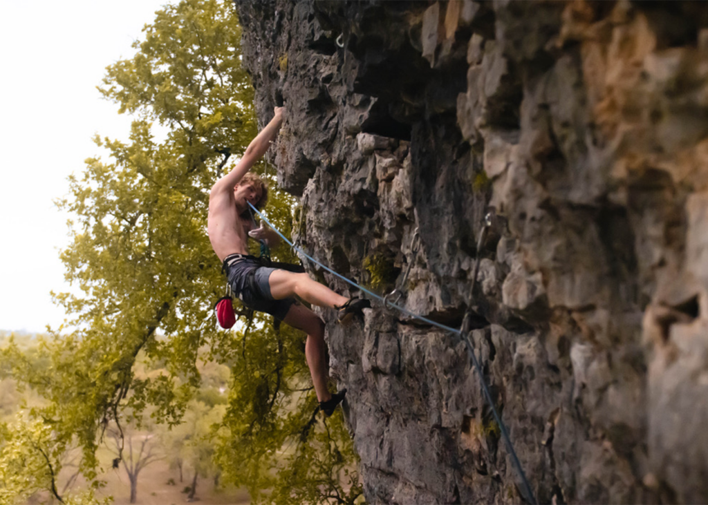 A person in shorts and no shirt climbs a steep, rocky cliff outdoors—secured by ropes—with trees and a blurred natural background visible, capturing the adventurous spirit of rock climbing Austin has to offer.