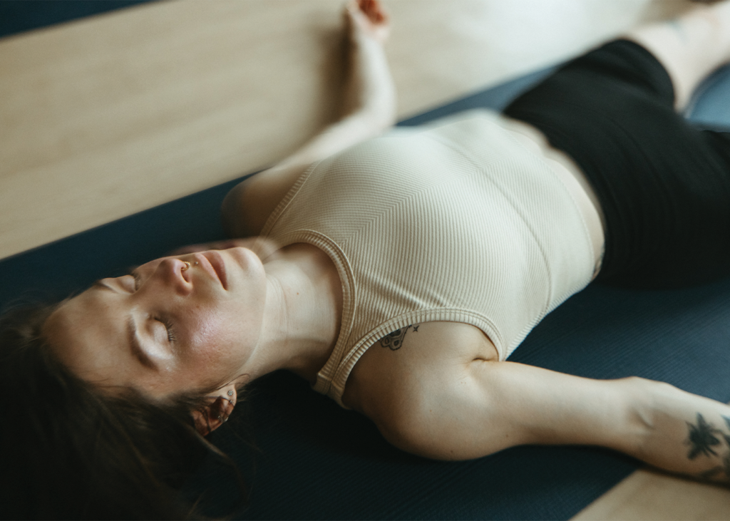 A person with closed eyes lies on a yoga mat, wearing a light tank top and dark shorts, appearing relaxed in the midst of their signature yoga practice. Their arms are resting by their sides, and they have a small tattoo on their upper arm.