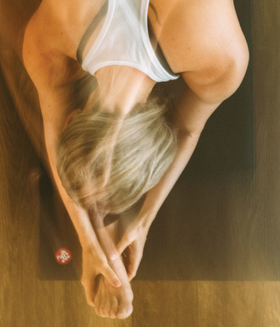 A person with blonde hair in a white tank top does a seated forward bend on a yoga mat, reaching for their foot during a Signature Yoga session. A red die is visible on the floor beside the mat.