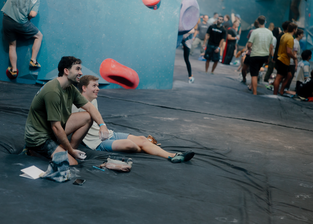 Two people sit on the floor at an indoor climbing gym in Austin, smiling and watching others climb. Climbing holds and gear are visible, with more people standing and climbing in the background—capturing the welcoming atmosphere that makes Austin rock climbing a great way to start rock climbing for beginners.