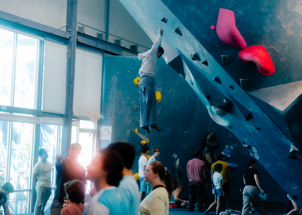A person in business attire hangs from an indoor rock climbing wall above a crowd of people. Bright climbing holds and natural light fill the busy Austin gym scene, offering inspiration for anyone curious about how to start rock climbing.