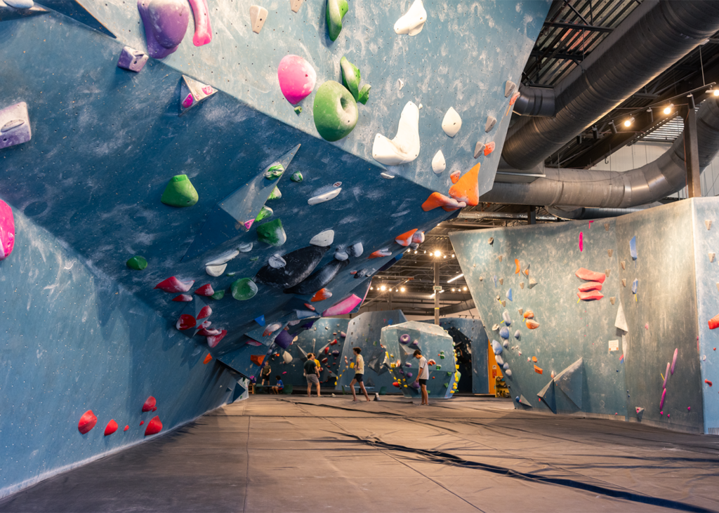 Indoor climbing gym with blue climbing walls, colorful climbing holds, and people bouldering. The space has padded floors and exposed ceilings with visible lighting and ventilation ducts—perfect for anyone looking to start rock climbing in the vibrant Austin rock climbing community.