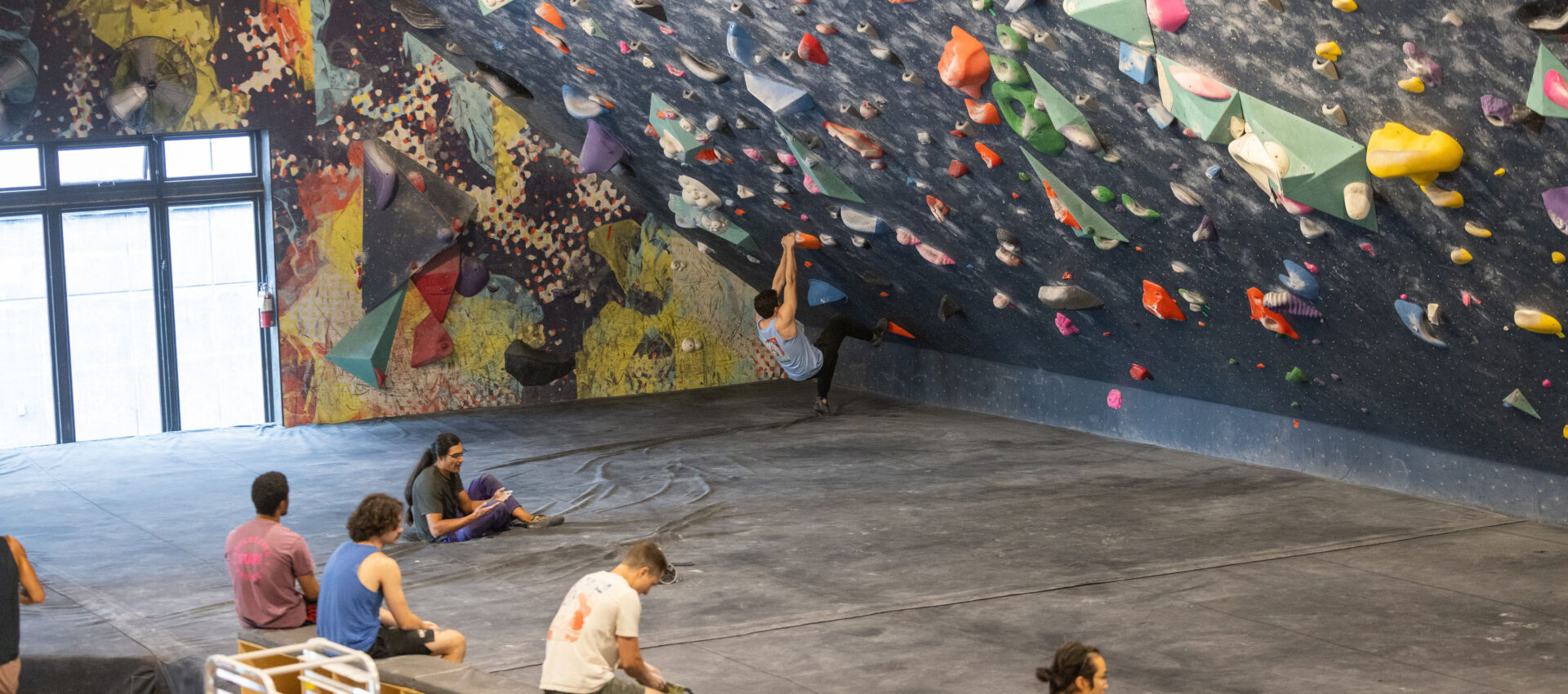 People sit and watch as a climber ascends a colorful indoor bouldering wall in a gym. The floor is padded, and sunlight shines in through large windows on the left, creating an inviting atmosphere—perfect for making the most of a summer membership.