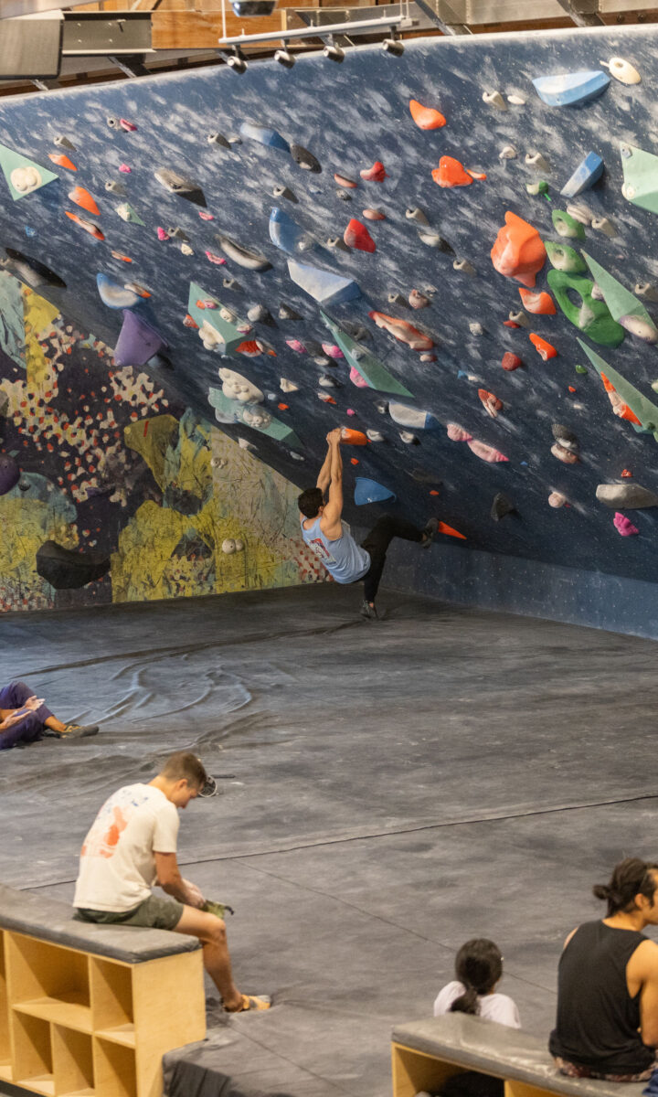 People sit and watch as a climber ascends a colorful indoor bouldering wall in a gym. The floor is padded, and sunlight shines in through large windows on the left, creating an inviting atmosphere—perfect for making the most of a summer membership.