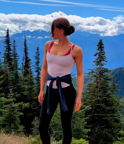 A woman in athletic wear stands outdoors, looking to the side, with poplar trees and mountains in the background under a partly cloudy sky near Seattle—a perfect setting for personal coaching amidst nature.