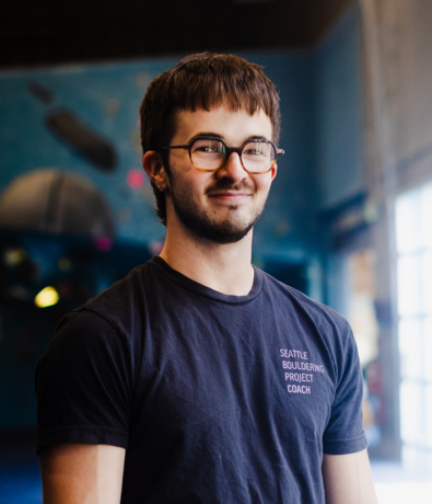 A young man with glasses, short brown hair, and a trimmed beard smiles while wearing a black Seattle Bouldering Project Coach shirt. The background features a blue indoor climbing gym in Seattle, highlighting his passion for personal coaching.