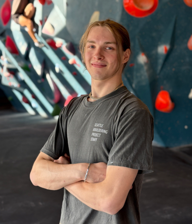 A person with long hair in a ponytail, wearing a grey Seattle Bouldering Project Staff shirt, stands with arms crossed in front of an indoor climbing wall with red holds, ready to offer personal coaching and guidance.