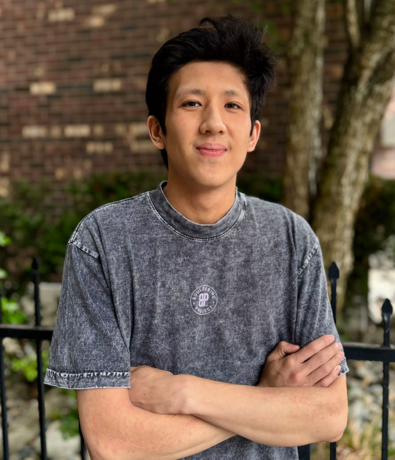 A young person with short dark hair stands outdoors with arms crossed, wearing a faded black T-shirt. Against the backdrop of a black iron fence, greenery, and a brick wall, they embody quiet confidence—an image that reflects the spirit of personal coaching in Seattle.