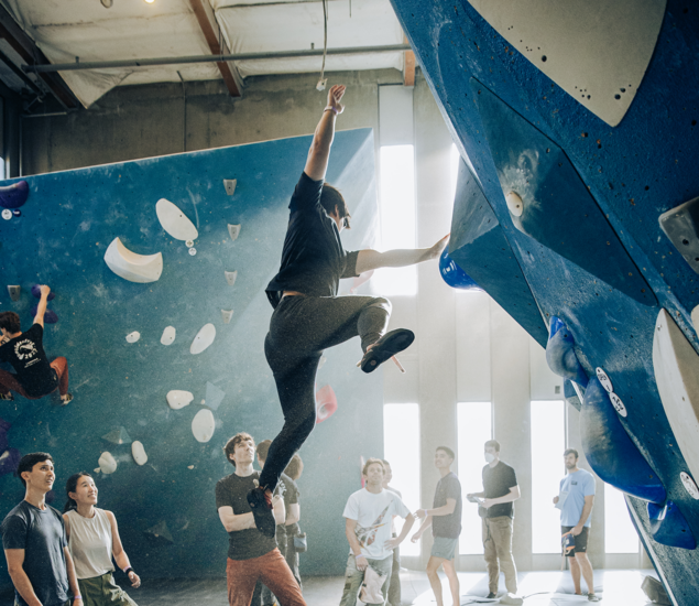 A person climbs an indoor bouldering wall in Seattle while others watch below. Sunlight streams through tall windows, and several climbers are visible on different parts of the blue wall, some benefiting from personal coaching as they improve their technique.