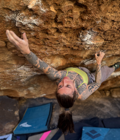 A person with tattoos climbs a steep, rocky surface near Poplar in Seattle, reaching upward with one hand. Crash pads are visible on the ground below, ready for a safe landing—perfect for anyone seeking personal coaching in bouldering technique.