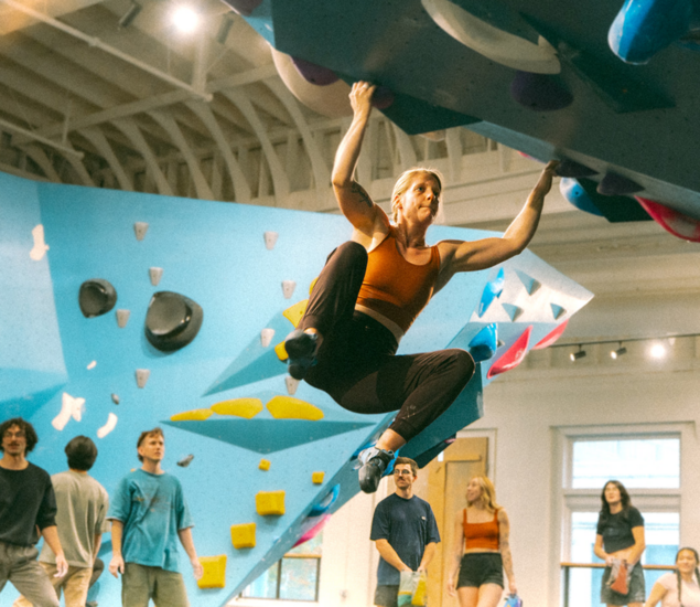 A person climbs an indoor bouldering wall, gripping holds on an overhang. Several people watch from below in a brightly lit climbing gym with blue walls and colorful holds—a hub for personal coaching in the heart of Seattle.