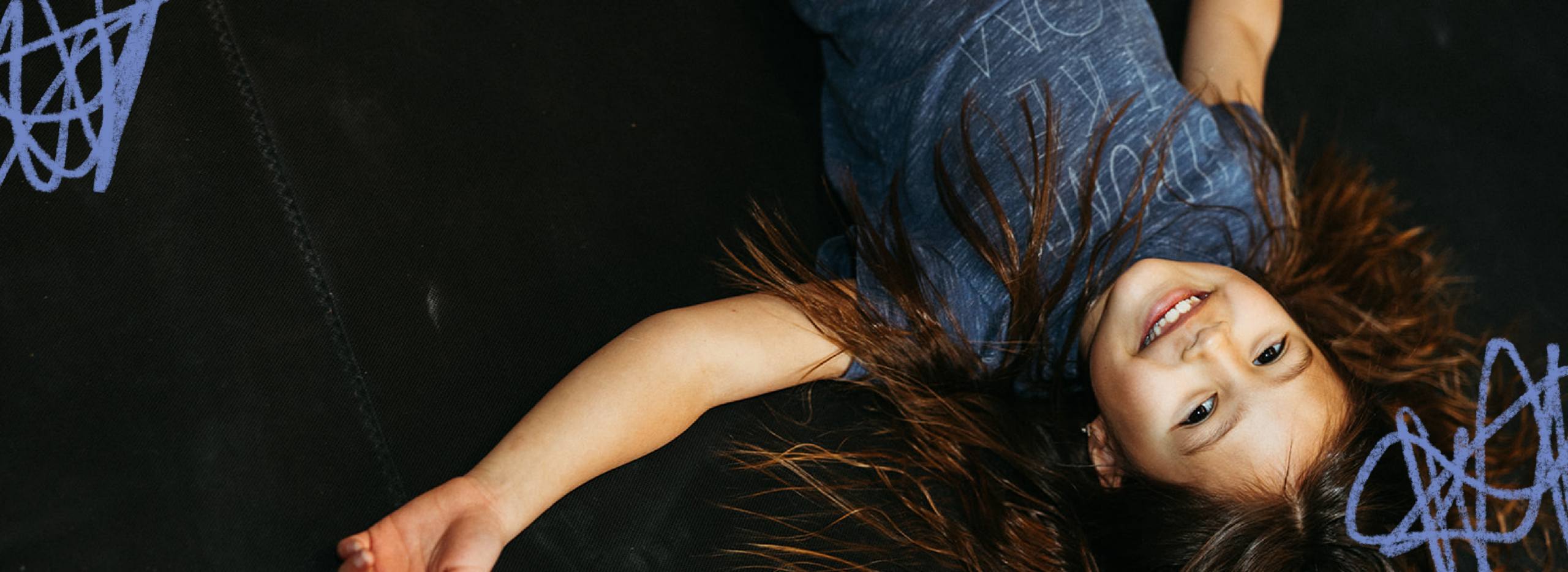A young girl with long hair lies on her back on a black bouldering gym surface, looking up and smiling.