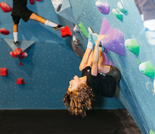 A child climbs a blue indoor rock wall during Austin youth programs, hanging upside down while gripping a large purple hold.