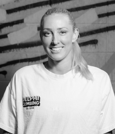A smiling woman wearing a white t-shirt with a Delphi Climbing Est. 2024 logo at a bouldering project.