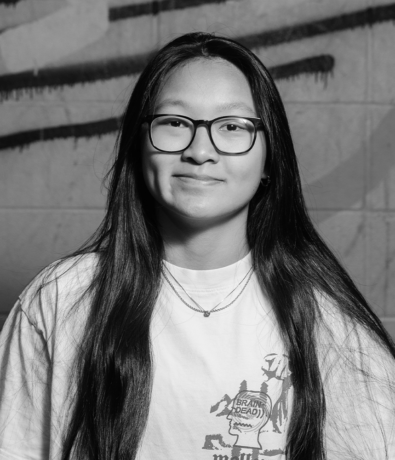 A young person with long dark hair and glasses wears a graphic T-shirt while posing against a graffiti-covered wall at a Boulderfest community climbing competition.