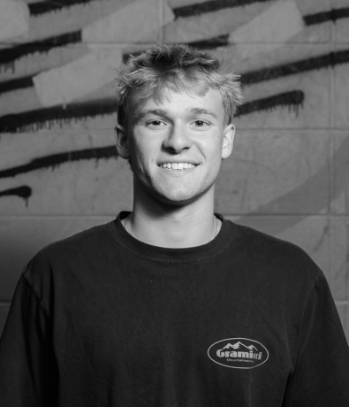 A young man smiling at the bouldering project in a black t-shirt with a Gramicci Mountaineering logo.